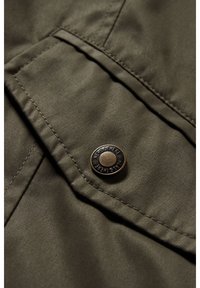 Tkanina w kolorze oliwkowej zieleni o gładkiej fakturze, z mosiężnym zatrzaskiem. Widoczne szycie wokół kieszeni i klapki.