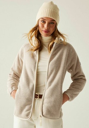 Mujer que lleva un gorro de punto beige, chaqueta con cremallera color beige claro, jersey de cuello alto crema y pantalones blanco hueso con cinturón marrón, de pie con las manos en los bolsillos.