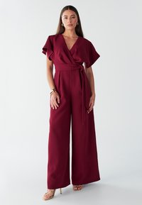 Burgundski kombinezon s kratkimi rahlo valovitimi rokavi, širokimi nogami, pasom in gladko teksturo. Kombiniran z zlatimi sandali s tankimi trakovi.