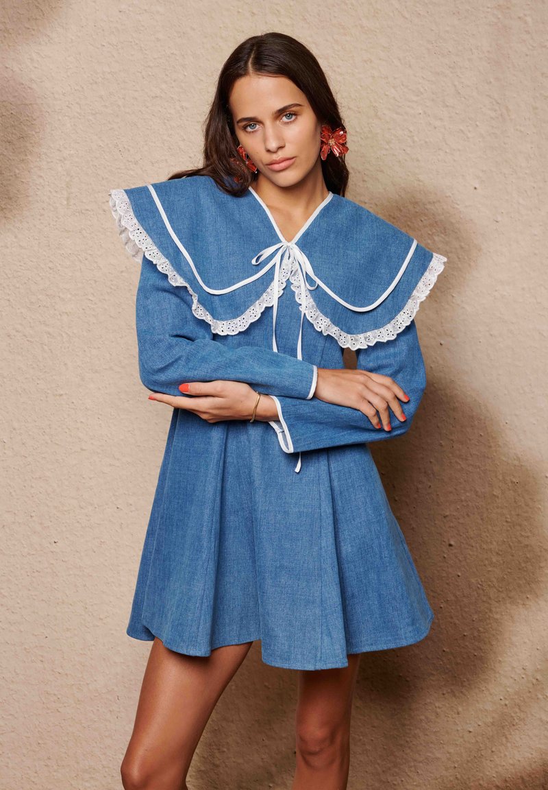 Τζιν φόρεμα με μεγάλο λευκό γιακά με δαντέλα και μακριά μανίκια. Το φόρεμα είναι φαρδύ με σφηκοφωλιά στη μέση και διακοσμητικό κορδόνι μπροστά.