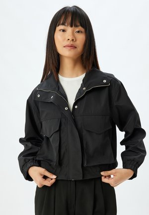 Chaqueta corta negra hecha de tejido suave, con cremallera frontal, dos grandes bolsillos de parche y puños elásticos. Diseño minimalista sin patrones.