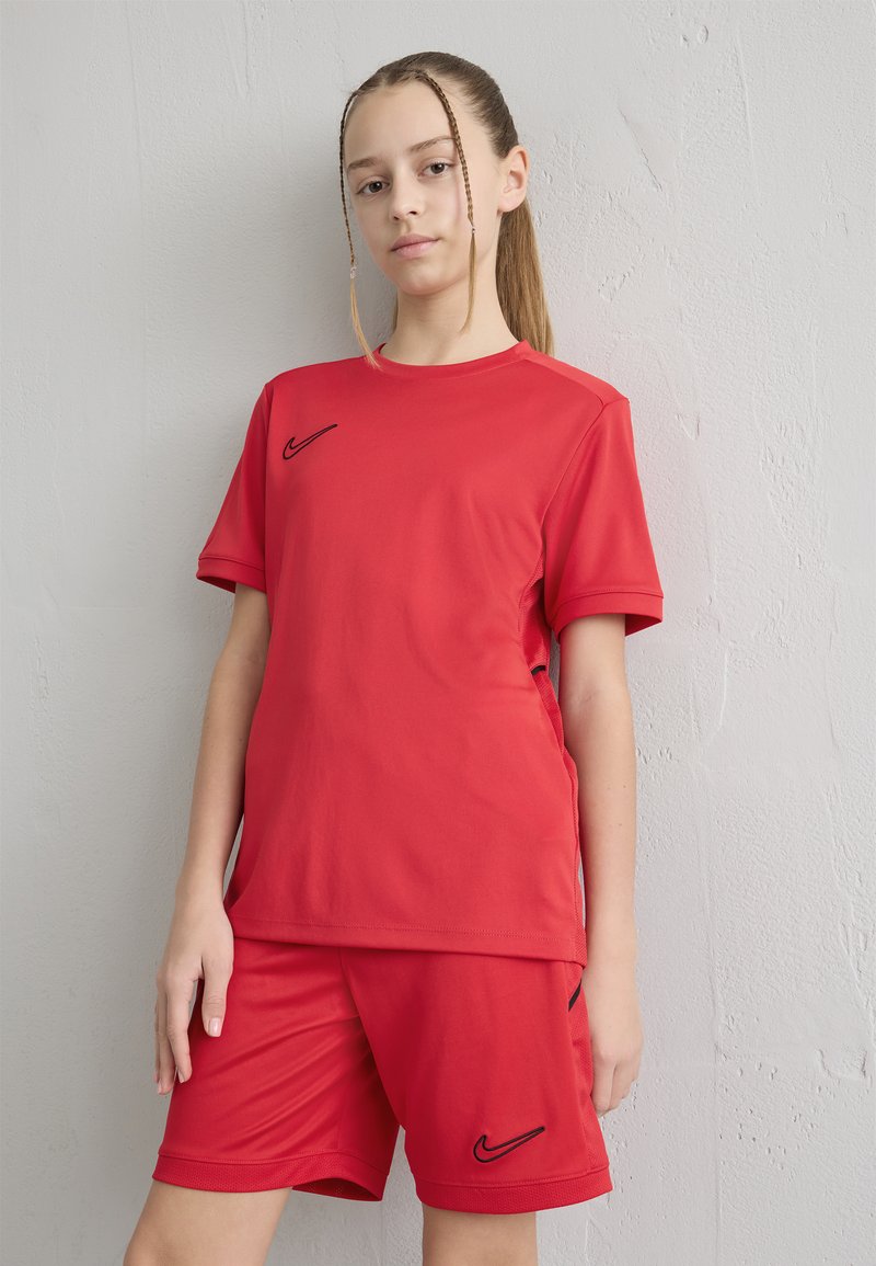 Adolescente con trenzas, vestida con ropa deportiva roja con logotipos negros de Nike, de pie contra una pared texturizada de color gris claro.