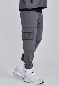 Man som bär grå cargojoggers, vita Nike-sneakers och vita strumpor, stående mot en enfärgad grå bakgrund.