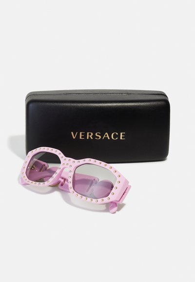 Roza prevelika sončna očala z zlatimi žebljički na okvirju, skupaj s črno torbico z zlatim logotipom Versace.