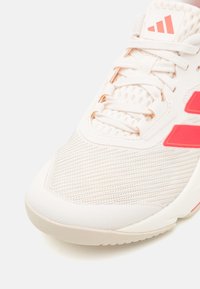 Białe buty sportowe z fakturowaną cholewką, z różowymi akcentami i trzema charakterystycznymi paskami. Posiadają zaokrąglony nosek i sznurowane zapięcie.