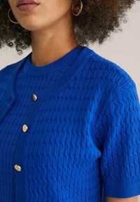 Cardigan en tricot bleu avec un motif texturé, un col rond et trois boutons dorés sur le devant. Les manches courtes achèvent le look.