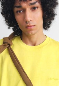 Camiseta de algodón amarilla con cuello redondo, que presenta una marca en relieve. Una banda de tela marrón cae sobre el hombro, añadiendo contraste.