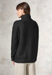 Schwarzer Strickpullover mit hohem Kragen, strukturiertem Stoff, langen Ärmeln und geripptem Saum. Das Model trägt hellfarbige Hosen.