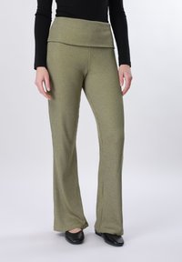 Γυναίκα που φοράει ελαιί πράσινο ριμπ παντελόνι φαρδιάς γραμμής με διπλωμένη μέση και μαύρα παπούτσια, στέκεται μπροστά σε απλό φόντο.