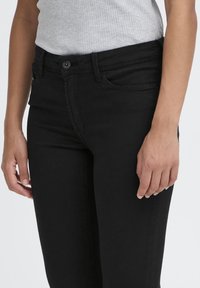 Personne portant un jean noir ajusté et un haut sans manches gris côtelé, debout avec les mains détendues le long du corps sur un fond uni.