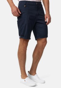 Shorts en coton bleu marine avec des ourlets repliés, présentant un bouton avant et deux poches latérales. Portés avec des chaussures blanches à enfiler.