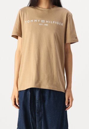 Beżowy T-shirt Tommy Hilfiger z krótkim rękawem i białym logo, noszony przez osobę w ciemnoniebieskiej spódnicy dżinsowej na jednolitym tle.