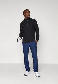 Man i svart tröja med kvartszip, blå byxor, vita sneakers och golfhandske, håller en golfklubba vilande på axeln och tittar åt sidan.