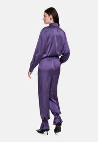 Purple satin two-piece outfit featuring a collared, button-up shirt and tapered pants with drawstring cuffs, adorned with subtle embellishments.