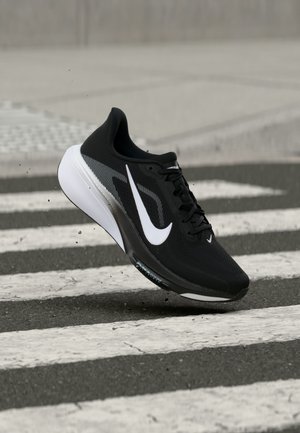 Zwart-witte Nike hardloopschoen zwevend boven een zebrapad in een stad met kleine stofdeeltjes rond de zool.