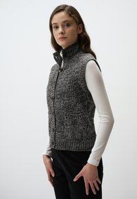 Gilet grigio a maglia con colletto alto, bottoni frontali e tasche laterali; indossato sopra una maglietta bianca a maniche lunghe con un motivo testurizzato.