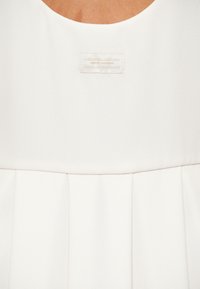 Λευκό αμάνικο τοπ με στρογγυλή λαιμόκοψη, πιεττέ χαμηλότερο τμήμα και μια μικρή ορθογώνια ετικέτα με την επιγραφή «Armani Exchange» κοντά στη λαιμόκοψη.