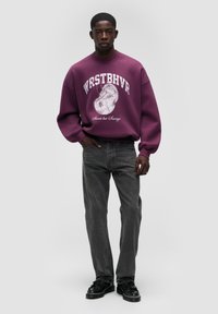 Felpa viola con grafica e testo bianco, abbinata a jeans grigio scuro e scarpe nere. Vestibilità comoda e texture morbida.