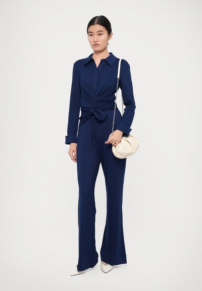 Navyblauwe jumpsuit met lange mouwen en een knoopdetail in de taille. Heeft een kraag en uitlopende broekspijpen. Draagt een crèmekleurige handtas.