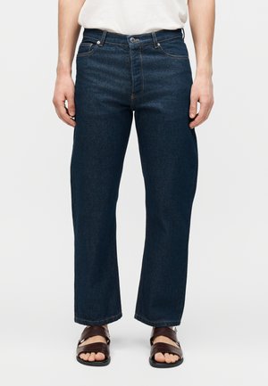 Hombre con vaqueros azul oscuro de corte recto, camisa blanca por dentro y sandalias marrones abiertas en los dedos, de pie frente a un fondo claro y liso.