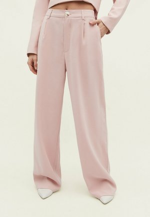 Hellrosa weit geschnittene Hosen mit glatter Textur, vorderen Falten und einem zentralen Knopfverschluss. Kombiniert mit spitzen weißen Schuhen.