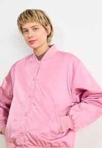 Ružový saténový bomber s vysokým golierom, patentkami a rebrovanými manžetami. Má postranné vrecká a hladkú, elegantnú textúru.