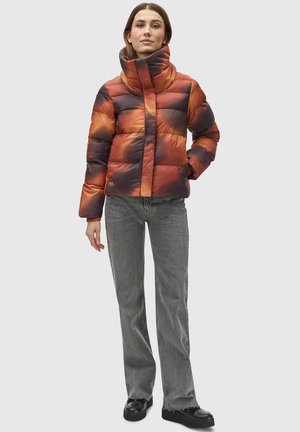 Pufferjack in een vervaagde oranje en zwarte kleur, met een hoge kraag, gewatteerd ontwerp en ritssluiting aan de voorkant. Combinatie met grijze broek en zwarte schoenen.