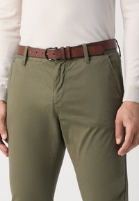 Pantalones de algodón verde oliva con una textura suave, combinados con un cinturón de cuero marrón que cuenta con una hebilla rectangular de plata y múltiples agujeros de ajuste.