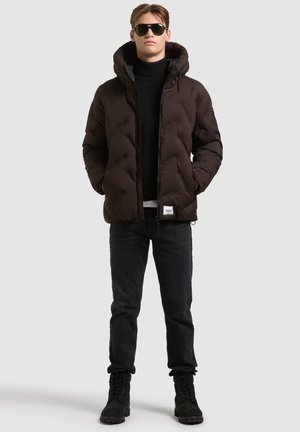 Homme portant une doudoune noire, un col roulé noir, un jean foncé, des bottes noires et des lunettes de soleil noires, debout devant un fond clair uni.