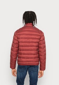 Rød pufferjakke med en skinnende, quiltet tekstur, med høj krave og metal hardware detaljer. Båret med mørkeblå jeans.