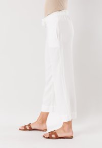 Pantaloni bianchi a gamba larga con cintura elastica e tasche laterali. Presentano una texture liscia e orli rifiniti. Abbinati a sandali bassi marroni.