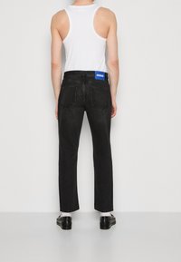 Persona con jeans negros y etiqueta azul "Hugo" en la parte trasera de la cintura, camiseta sin mangas blanca, calcetines blancos y mocasines negros, mirando de espaldas a la cámara.