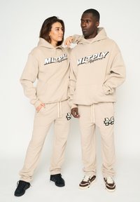 Felpe e pantaloni della tuta beige con una consistenza morbida, caratterizzati da accenti del logo in nero. Gli abiti hanno una vestibilità comoda e dettagli con coulisse.