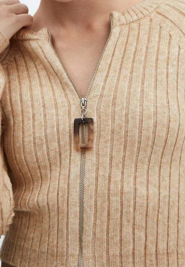 Pull côtelé beige avec une fermeture éclair argentée et un tire-zip carré en écaille de tortue porté par une personne avec une main visible près du col.