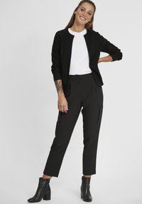 Cardigan noir avec motif texturé, T-shirt blanc et pantalon noir ajusté. Associé avec des bottines. Fond neutre.