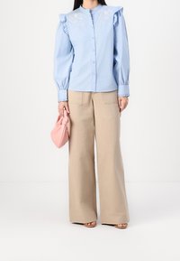 Camicia azzurro chiaro con maniche arricciate e ricami floreali, abbinata a pantaloni ampi beige e una borsa a tracolla rosa con nodo.