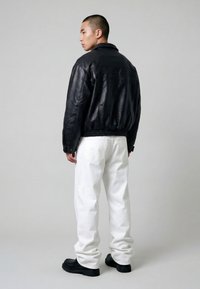 Mand iført sort læderjakke, hvide bukser og sorte sko, stående sidelæns mod en ensfarvet grå baggrund.