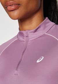 Lähikuva henkilöstä, jolla on yllä violetti vetoketjullinen urheiluvaruste, jossa rinnan kohdalla on valkoinen Asics-logo.