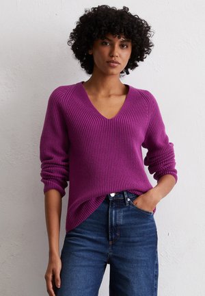 Lebhaftes lila Strickpullover mit V-Ausschnitt, geripptem Muster und lockerer Passform. Mit hochgerollten Ärmeln und asymmetrischem Saum, kombiniert mit dunklen Jeans.