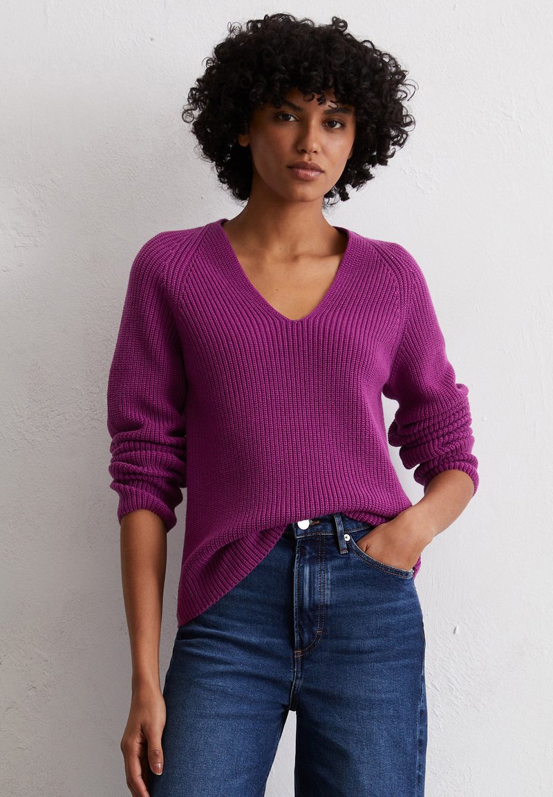 Lebhaftes lila Strickpullover mit V-Ausschnitt, geripptem Muster und lockerer Passform. Mit hochgerollten Ärmeln und asymmetrischem Saum, kombiniert mit dunklen Jeans.