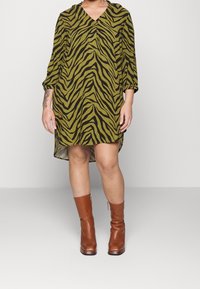 Zielona sukienka w tygrysie paski z dekoltem w kształcie V i miękkiego materiału, z długimi rękawami i asymetrycznym hemem. Noszona z brązowymi skórzanymi botkami.