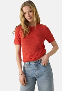 Pull rouge tricoté à manches courtes avec un motif texturé et des accents de bouton sur l'épaule, associé à un jean taille haute bleu clair.
