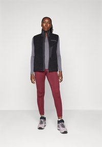 Black fleece vest with high collar, paired with a gray long-sleeve top and burgundy leggings. Model wears gray and black athletic shoes.