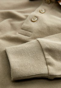 Beige Poloshirt mit Kragen aus strukturiertem Stoff, gerippten Bündchen und Knopfverschluss, der eine glatte und gleichmäßige Oberfläche zeigt.