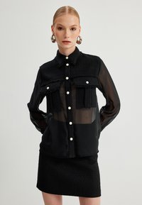 Schwarze, knöpfbare Bluse mit einer strukturierten, ausgefransten oberen Schicht; transparente Ärmel; zwei Brusttaschen; Perlmuttknöpfe; kombiniert mit einem passenden Rock.