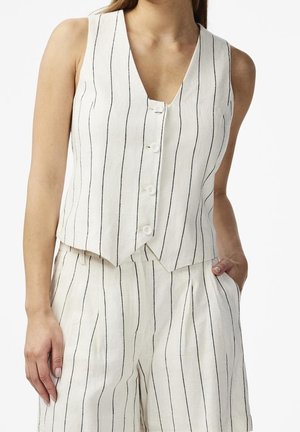 Kobieta ubrana w białą bezrękawnik i pasujące szerokie spodnie z cienkimi pionowymi czarnymi paskami, jedna ręka w kieszeni, na białym tle.