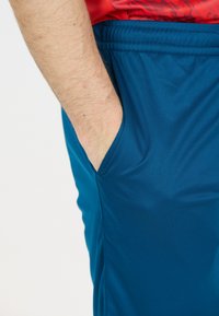 En vuxens hand delvis i höger ficka på blå stretchbyxor med resårmidja och en röd skjorta ovanför.