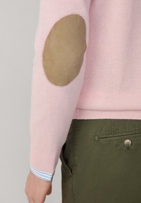 Manica di maglione rosa chiaro con toppa ovale beige sul gomito, indossata sopra una camicia a righe blu e bianche, abbinata a pantaloni verde oliva con tasca sul retro con bottone.