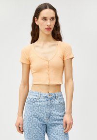 Kurzärmelige, cropped Strickjacke aus geripptem, hellorangefarbenem Stoff mit V-Ausschnitt und fünf Knopfverschluss. Tragen mit hochgeschnittenen gemusterten Jeans.