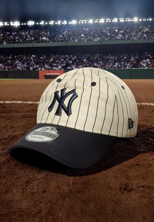 Beige baseball cap with navy accents, featuring vertical pinstripes and an embroidered logo on the front. Adjustable sizing.